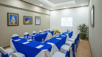 A conference room with long tables, white chairs, and blue tablecloths. There are framed pictures on the wall and a projector screen displaying a logo.