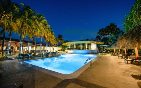 A swimming pool with lounge chairs and palm trees, surrounded by a building with a thatched roof.