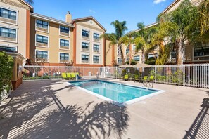 A swimming pool with a white fence, surrounded by palm trees and a building with multiple windows and balconies.