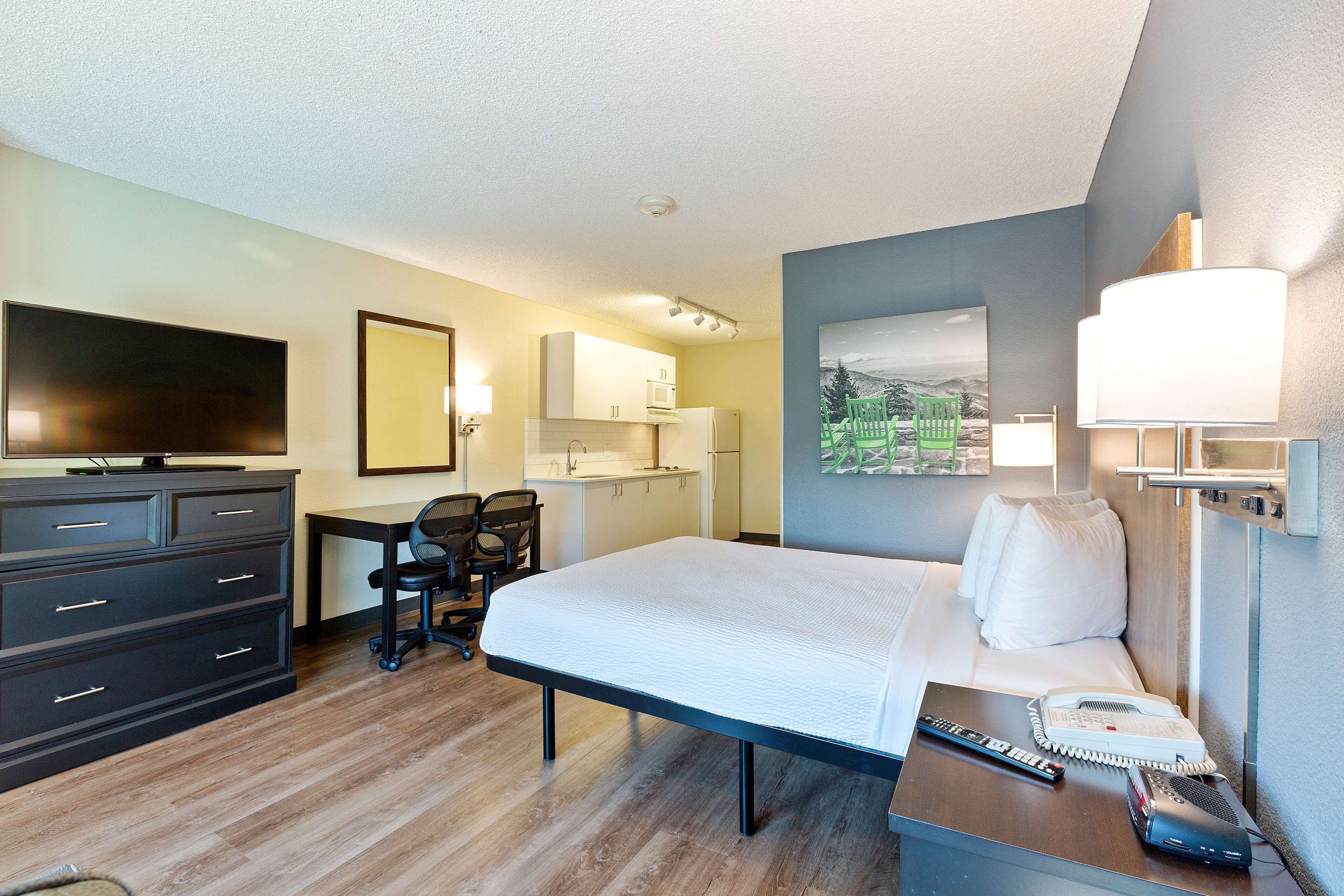 A hotel room with a bed, desk, chair, TV, and kitchenette.