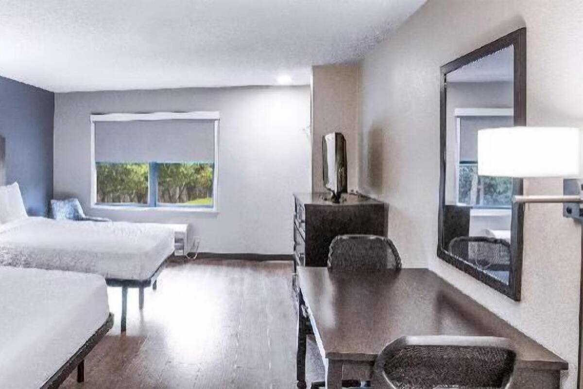 A hotel room with two beds, a desk, a chair, a window, and a mirror.