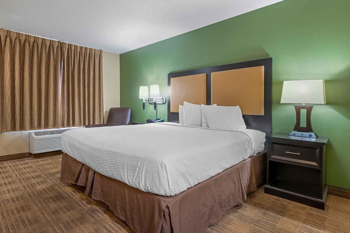 A hotel room with a large bed, a desk, a chair, a lamp, and a wall-mounted heater.