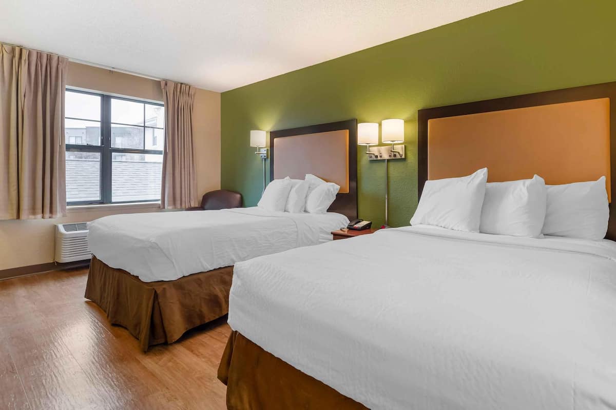 A hotel room with two beds, a window with curtains, a wall-mounted lamp, and an air conditioning unit.