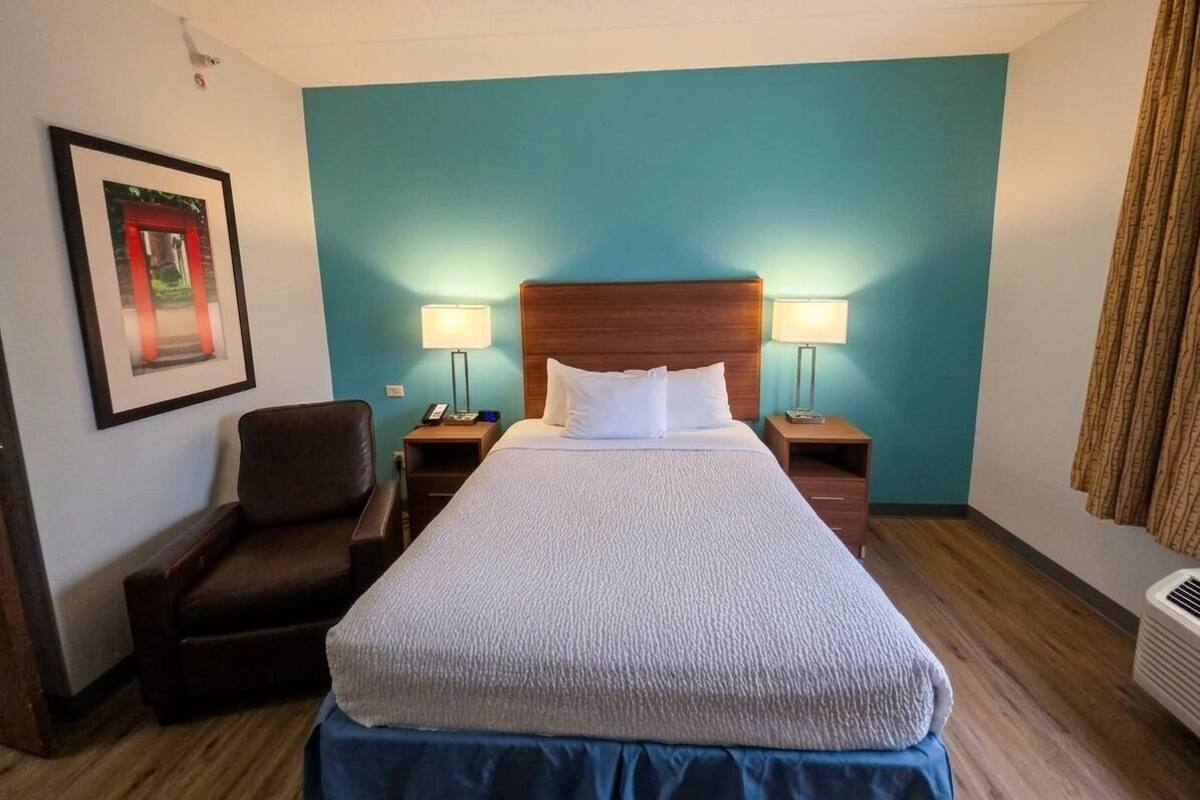 A hotel room with a bed, two bedside lamps, a chair, a framed picture on the wall, and a window with curtains.