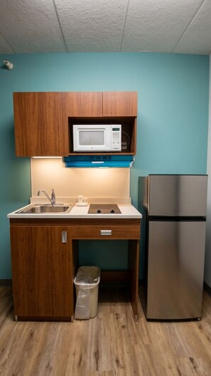 A compact kitchenette with wooden cabinets, a microwave, a stainless steel refrigerator, a sink, and a trash bin. - MainStay Suites Chicago Hillside (Hillside)