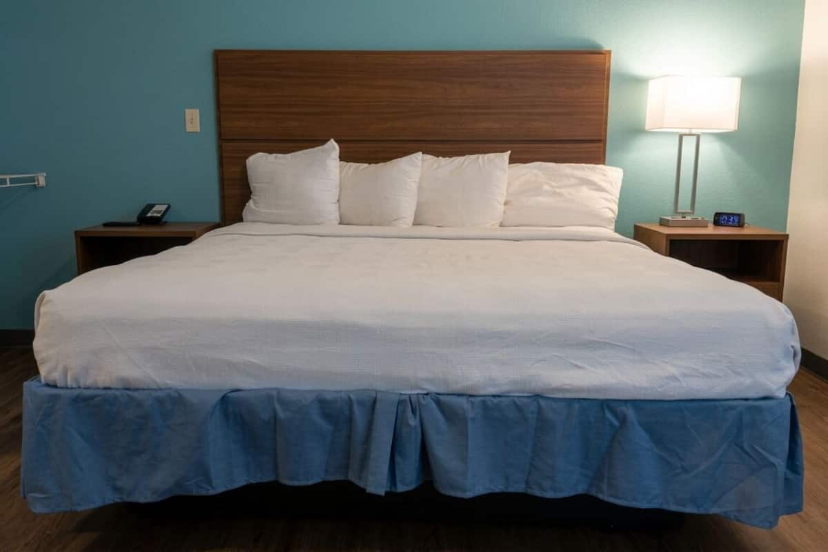 A hotel room with a large bed, wooden headboard, white bedding, a blue skirted bed base, a bedside table with a lamp, and a nightstand with a phone.
