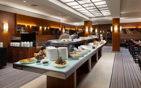 A buffet area with a variety of food items, including fruits, vegetables, and prepared dishes, set in a modern hotel with wooden accents and a high ceiling.