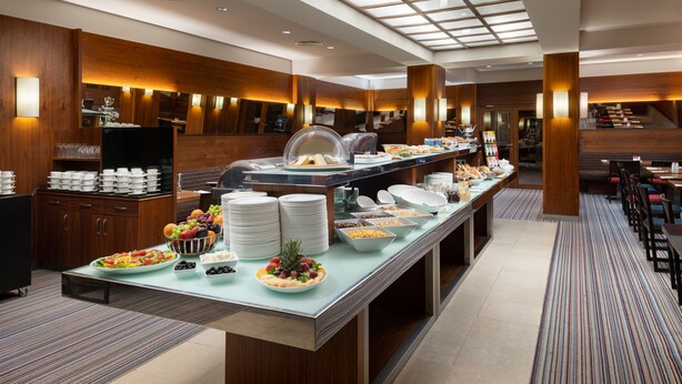 Un espace buffet proposant une grande variété d’aliments, notamment des fruits, des légumes et des plats préparés, situé dans un hôtel moderne agrémenté d’éléments en bois et doté d’un plafond haut.