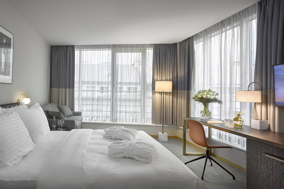 Quarto de hotel com cama, escrivaninha, cadeira, televisão e janelas amplas com cortinas.