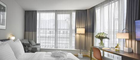 Habitación de hotel con cama, escritorio con silla, televisor y ventanas grandes con cortinas.
