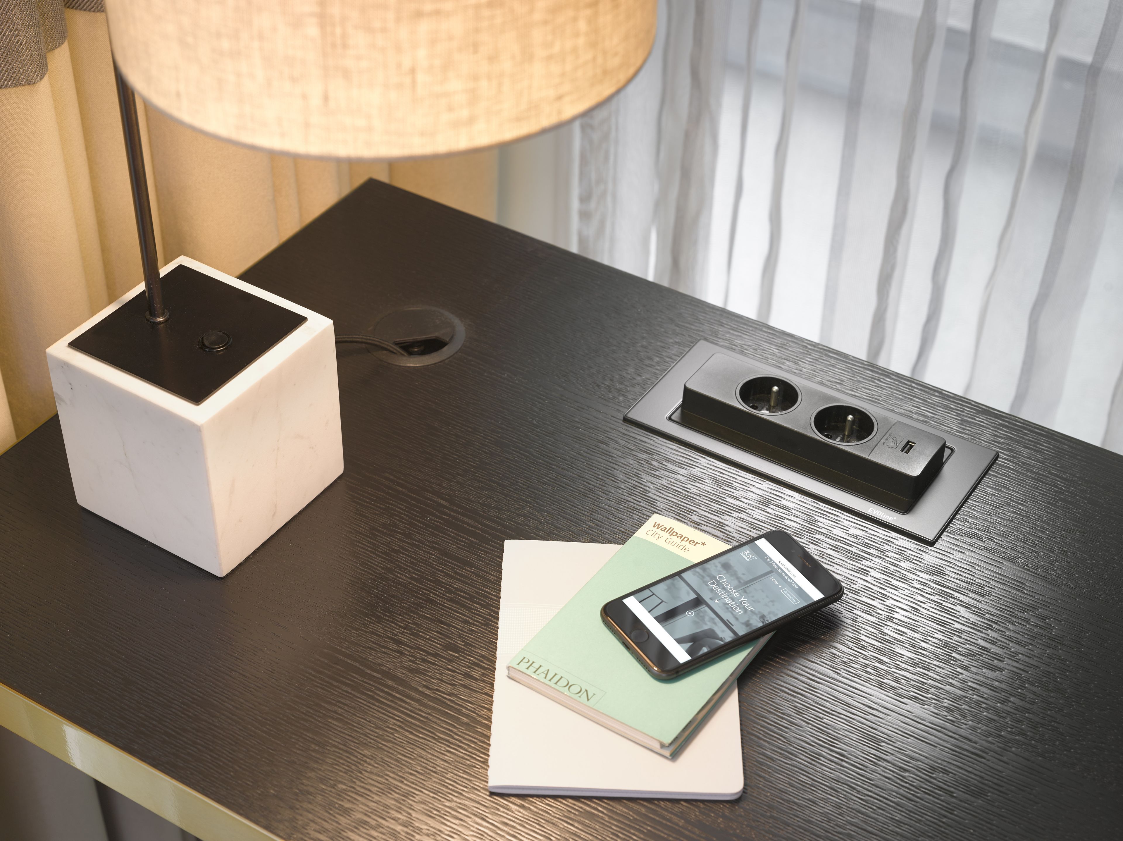 A hotel room with a desk featuring a lamp, a power strip, a phone, and a guidebook.
