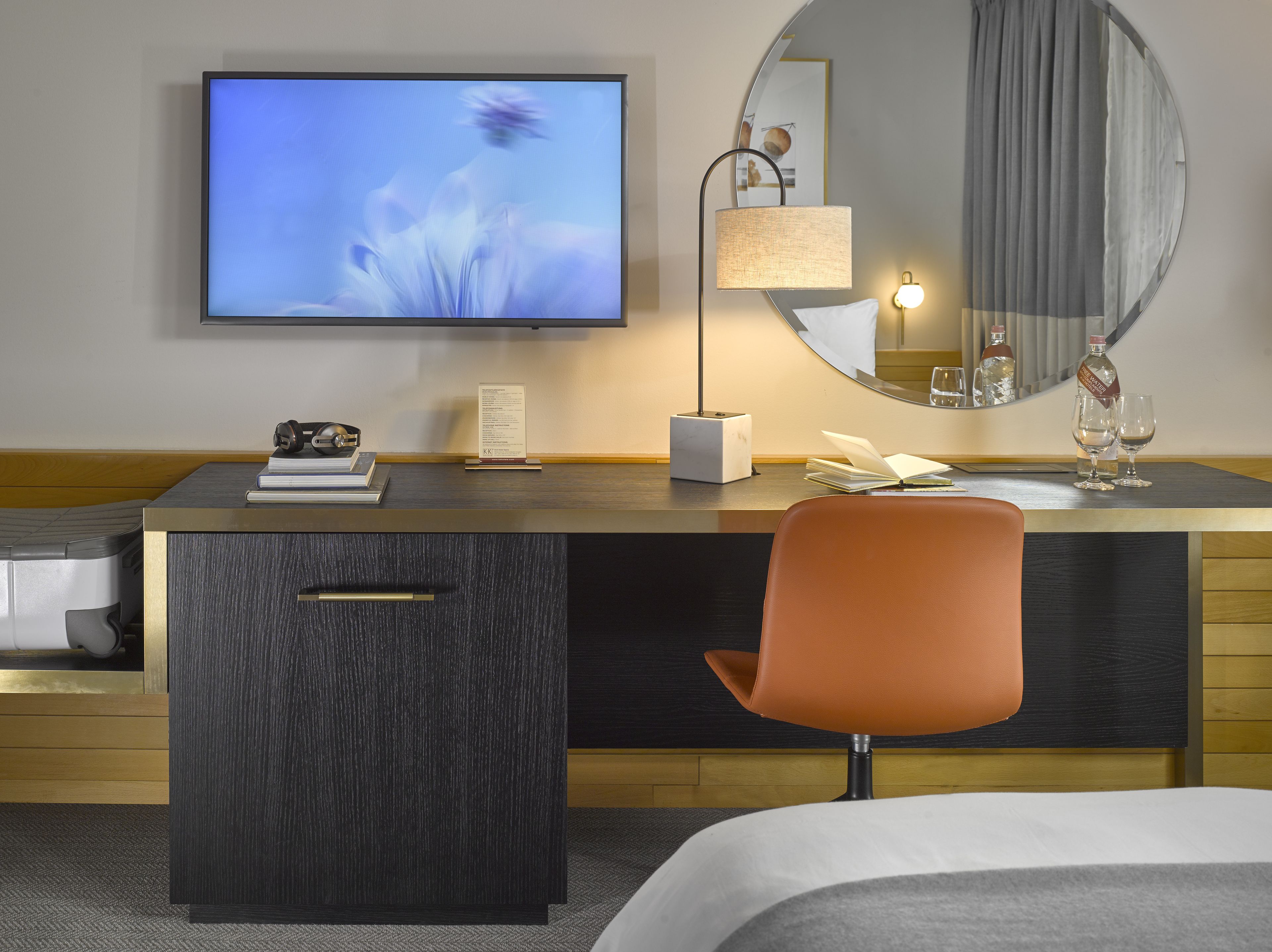 A hotel room with a flat-screen TV, a desk with a lamp, a chair, a bed, and a large mirror.