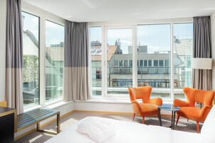 Camera d'albergo con un'ampia finestra, due poltrone arancioni, un tavolino, un letto e una panca.