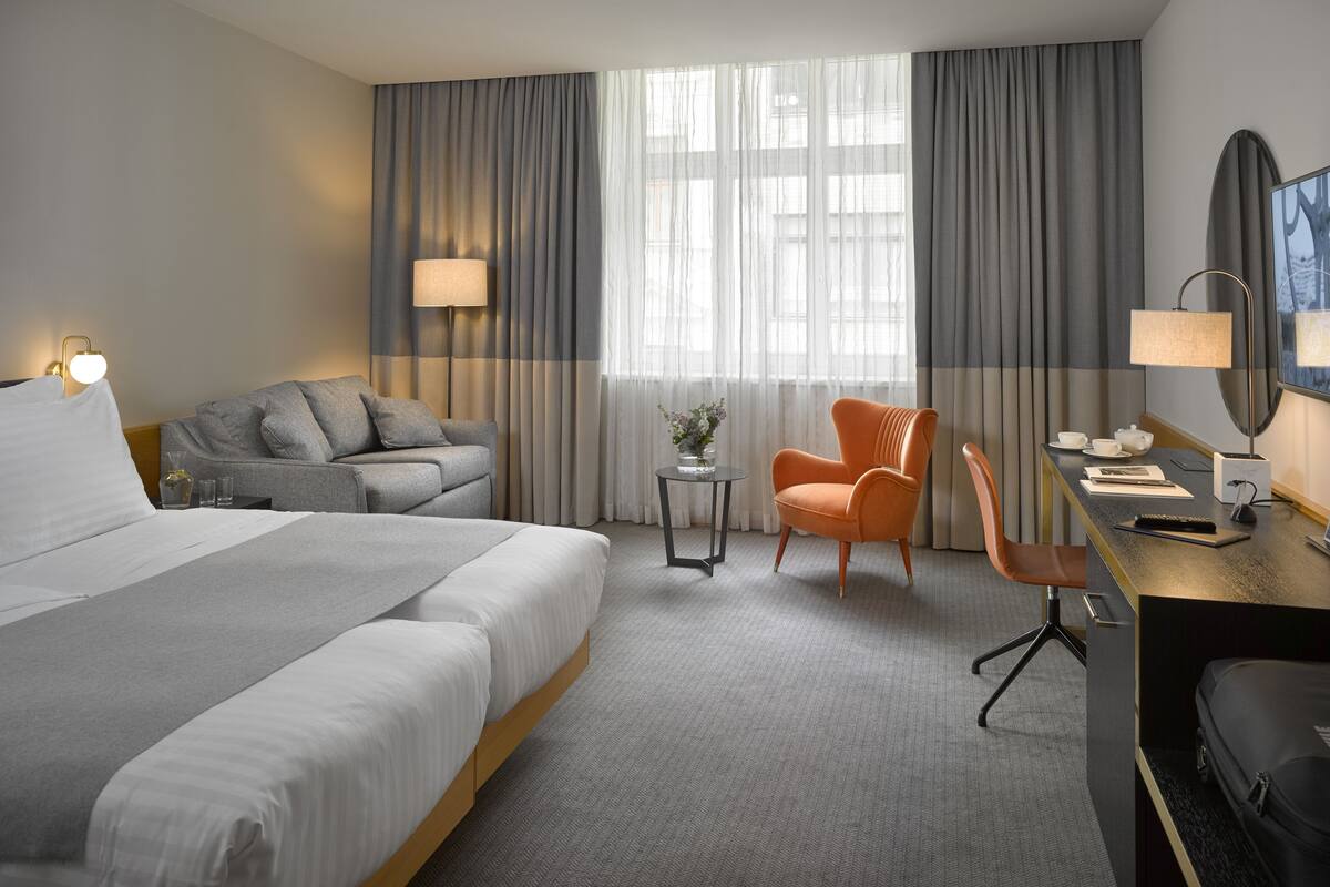 Quarto de hotel com uma cama grande, um sofá cinza, uma poltrona laranja, uma escrivaninha com abajur e uma janela com cortinas.