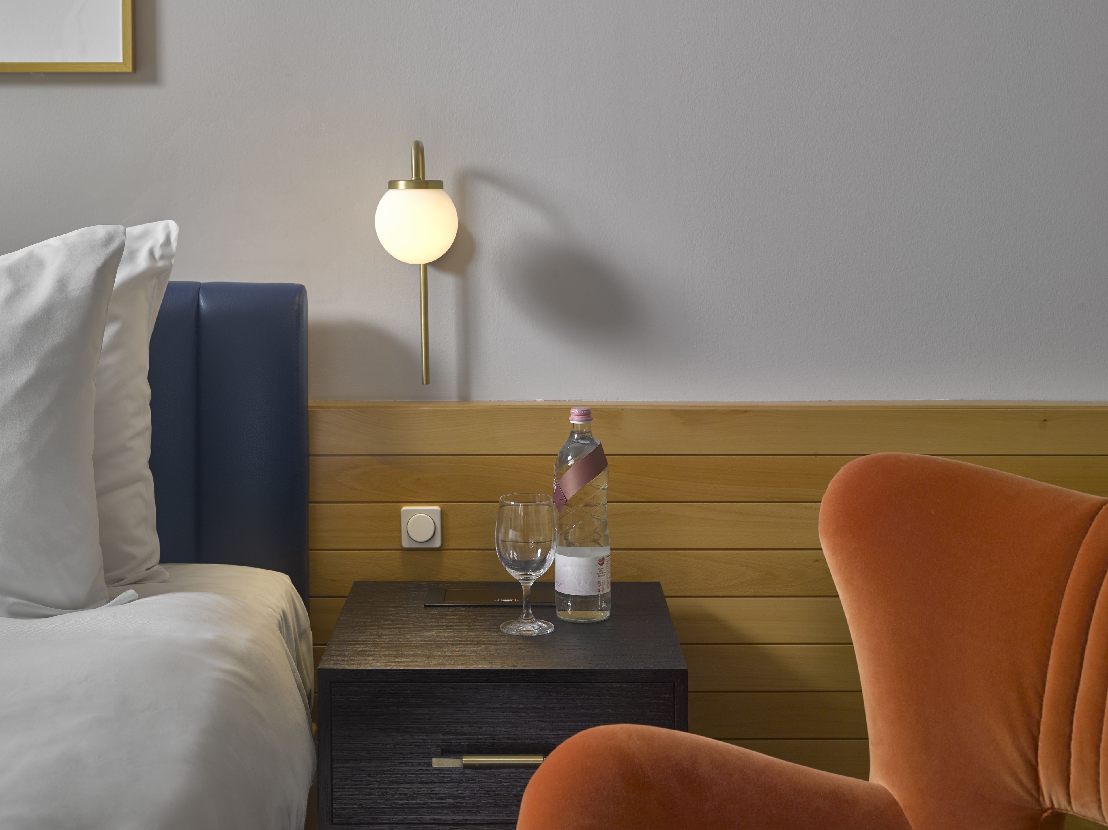 A hotel room with a bed, a nightstand, a lamp, a bottle of water, and a glass.