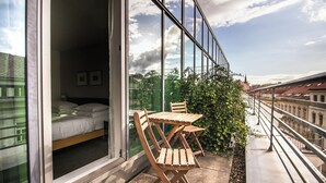 Un balcon aménagé avec un lit, des chaises en bois et une table, d’où l’on peut admirer un paysage urbain avec ses bâtiments et un ciel dégagé.