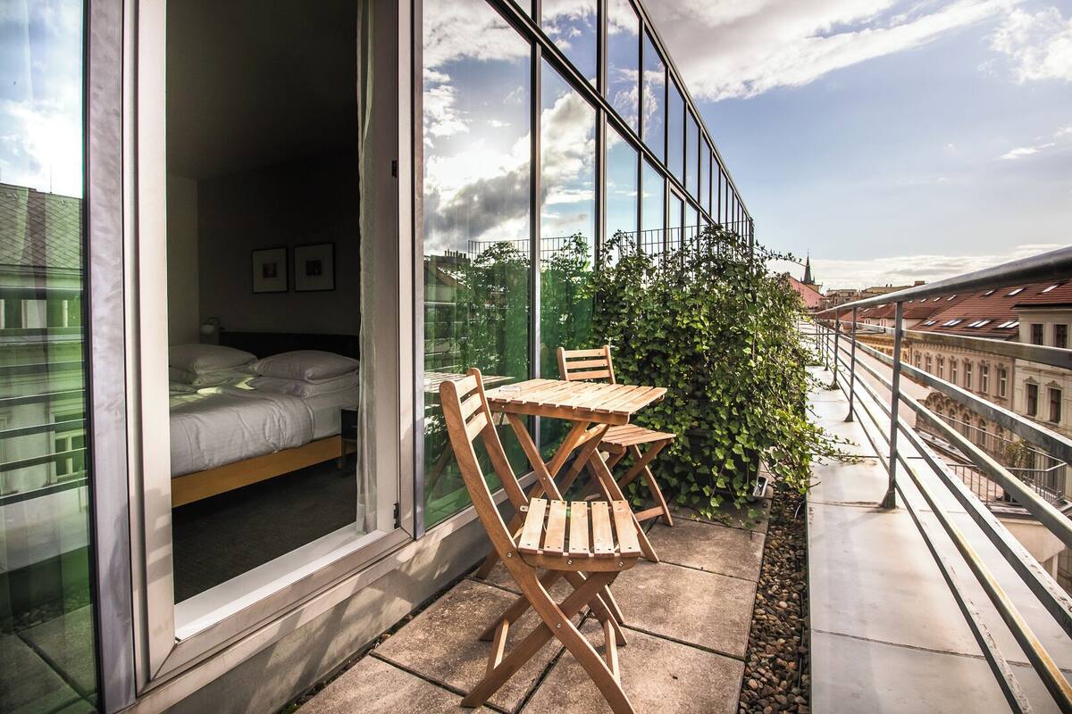 Varanda com cama, cadeiras de madeira e uma mesa, com vista para um horizonte urbano com edifícios e céu claro.