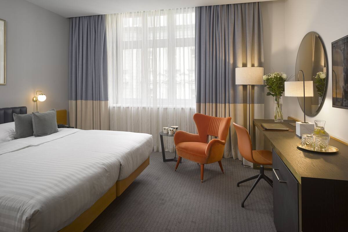 Quarto de hotel com uma cama grande, duas cadeiras, uma escrivaninha com luminária, um vaso com flores e um espelho amplo.