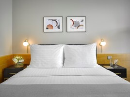 Uma cama bem arrumada com lençóis brancos, duas mesas de cabeceira com abajures, e duas pinturas abstratas emolduradas na parede.