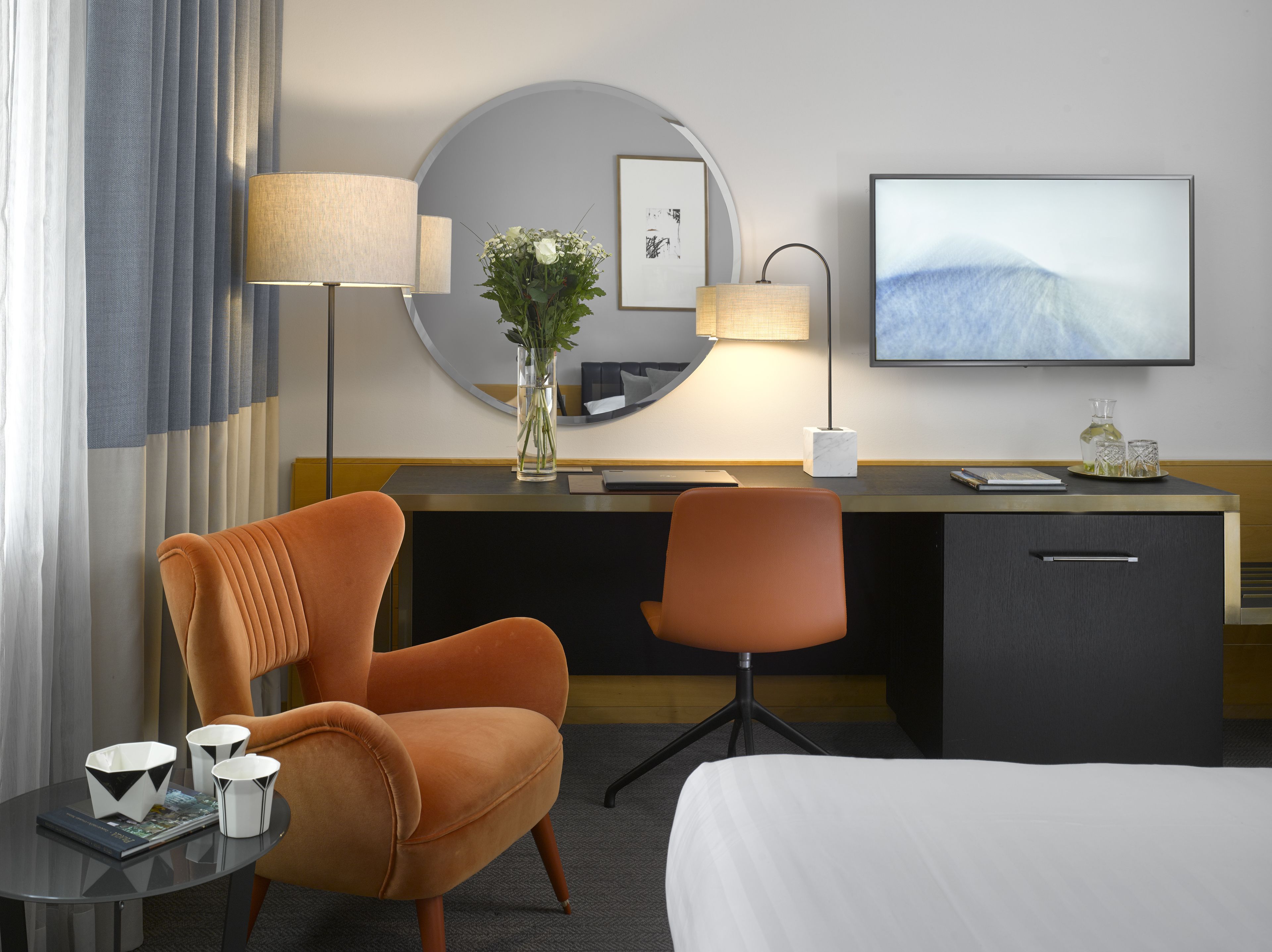 A hotel room with a bed, two chairs, a desk with a lamp, a vase with flowers, a framed picture, and a TV.