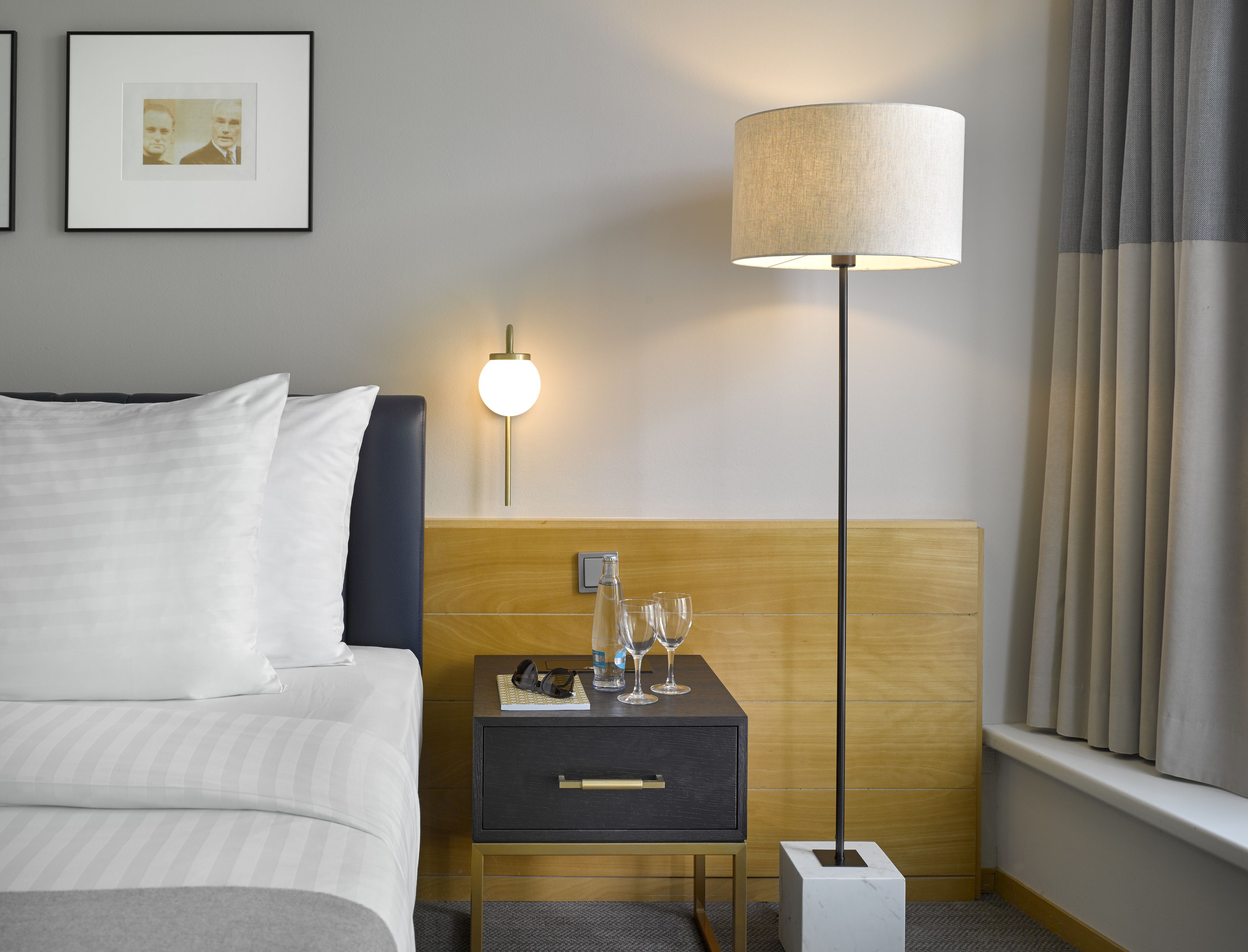 Een hotelkamer met een bed, nachtkastje met een lamp, een klein ladeblok en een raam met gordijnen.