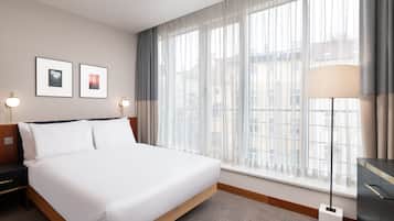 Quarto de hotel com uma cama grande, uma mesa de cabeceira, uma luminária e uma janela com cortinas.