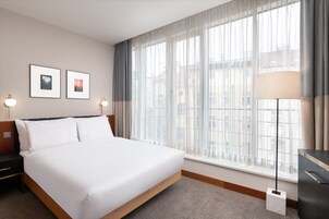 Quarto de hotel com uma cama grande, uma mesa de cabeceira, uma luminária e uma janela com cortinas.
