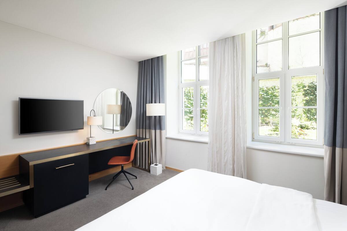 Quarto de hotel com cama, escrivaninha com cadeira, televisão, espelho e janela com cortinas.