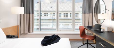 A hotel room with a bed, a desk, a chair, a lamp, and a window with curtains.