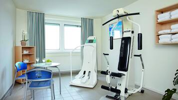 Un gimnasio equipado con cinta de correr, elíptica y máquinas de pesas. Hay una silla, una mesita con un vaso y un estante con toallas.