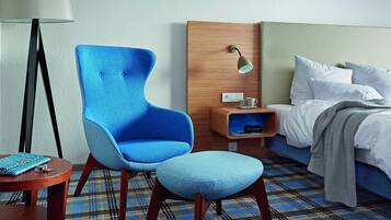A hotel room with a blue armchair, a bedside table with a lamp, a bed with white linens, and a plaid-patterned carpet.