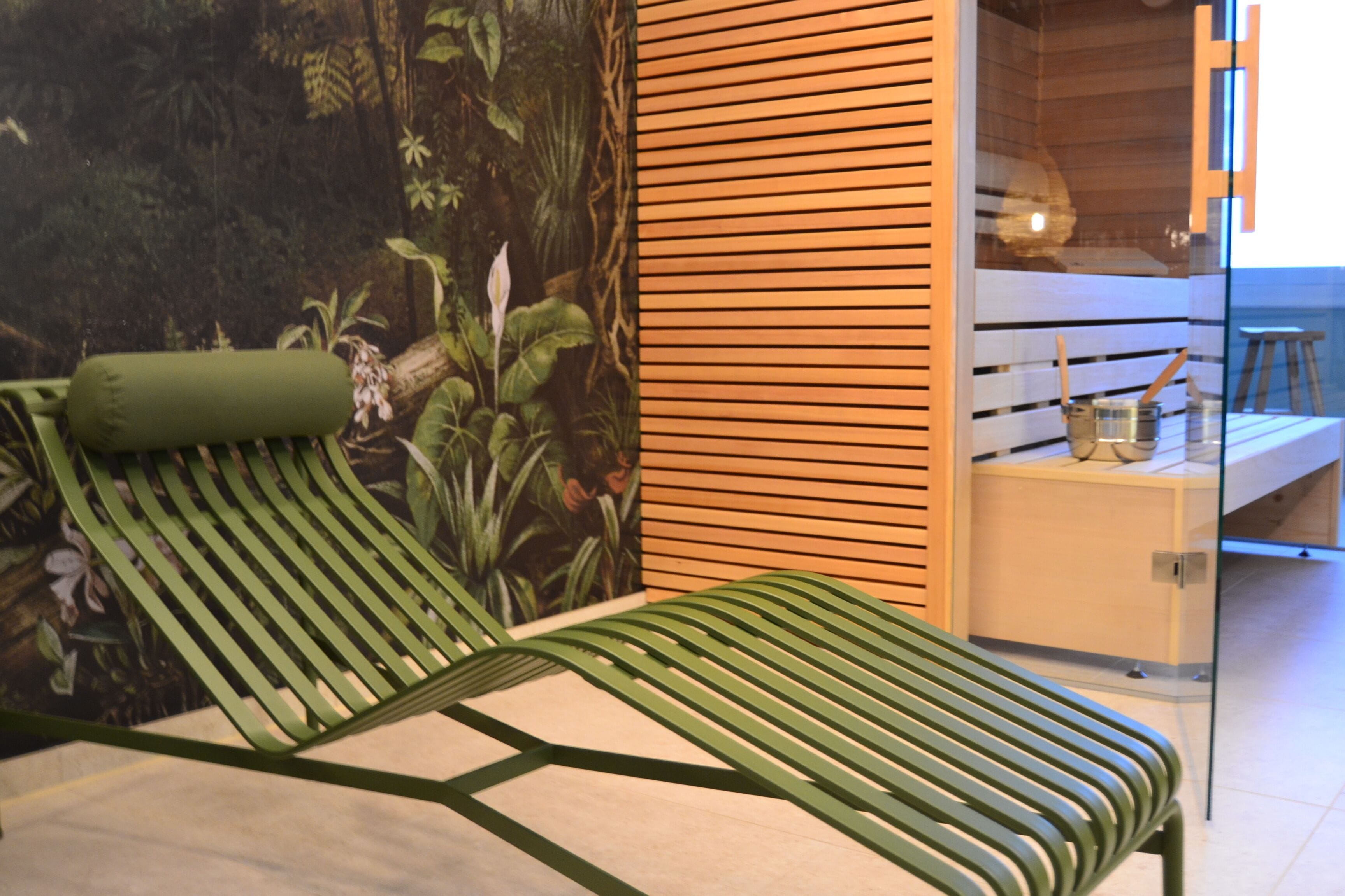 Ein grüner Loungesessel mit Kissen, eine Wand aus Holzlamellen und ein Glastrennfeld mit einem kleinen Hocker und einem Metallbehälter auf einer Theke.