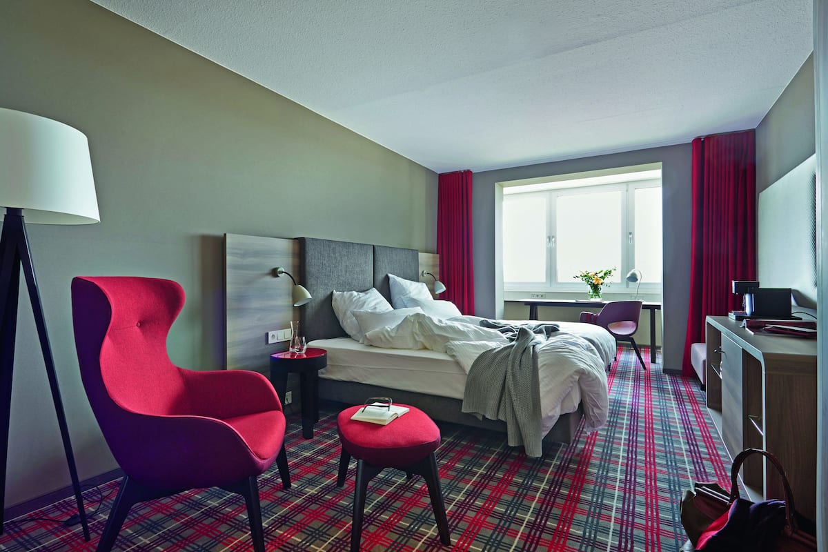 A hotel room with a bed, a red armchair, a small table, a desk, and a window with curtains.