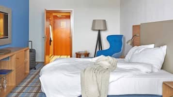 A hotel room with a bed, a desk, a chair, a lamp, and a door.