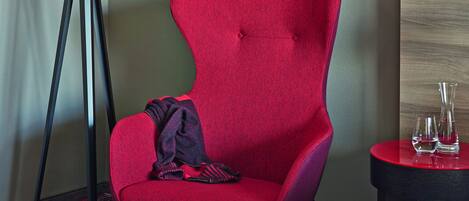 Habitación de hotel con un sillón tapizado rojo, una mesita redonda con una revista, una lámpara de pie y una mesita de noche con vasos.