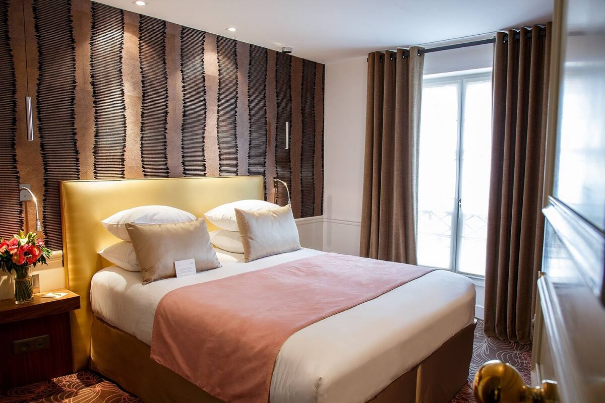 A hotel room with a large bed, a headboard, a bedside table with a lamp, and a window with curtains.
