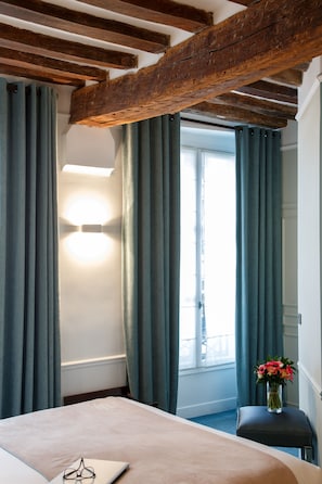 Une chambre d’hôtel avec un lit, une fenêtre avec des rideaux, une lampe de chevet et un vase rempli de fleurs sur une petite table.