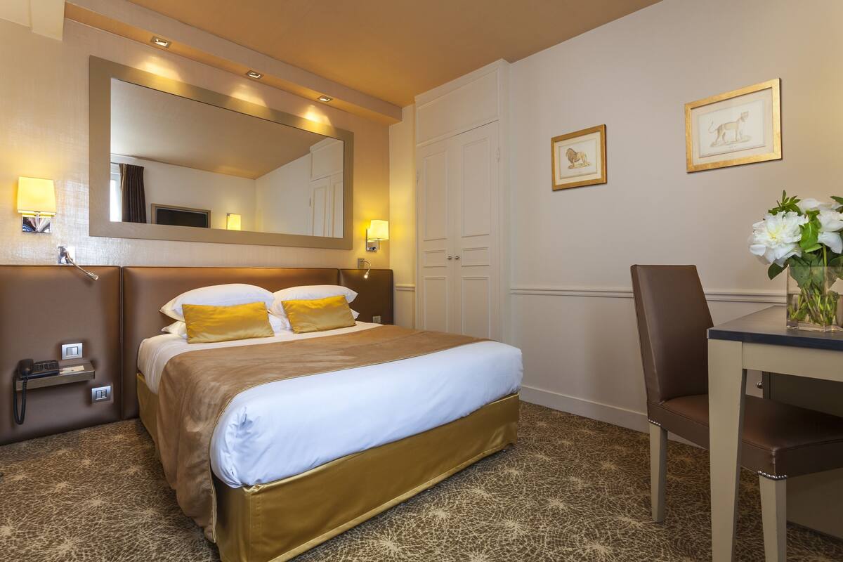 A hotel room with a large bed, a desk with a chair, a mirror, and framed pictures on the wall.