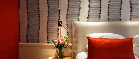 Una habitación de hotel con cama, mesita de noche, un florero con flores y una ventana con persianas.