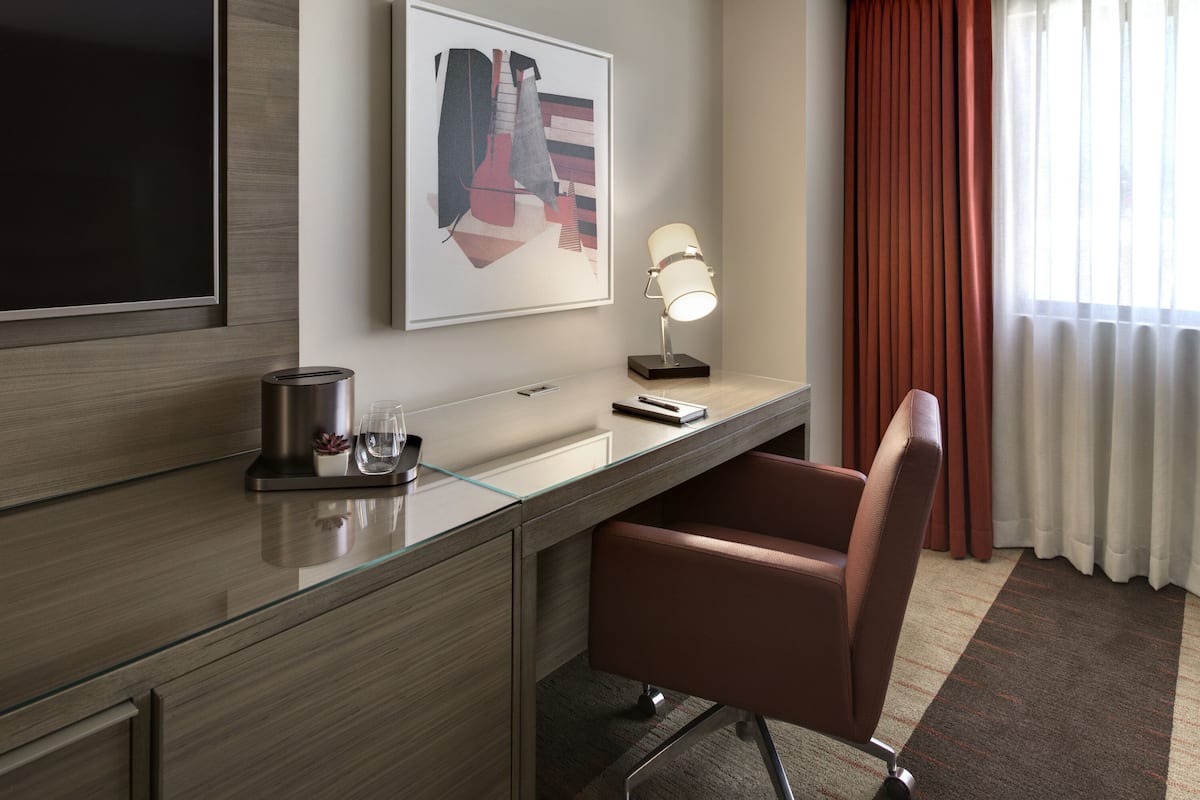 A modern hotel room with a wooden desk, a flat-screen TV, a framed abstract artwork, a desk lamp, and a brown leather chair.