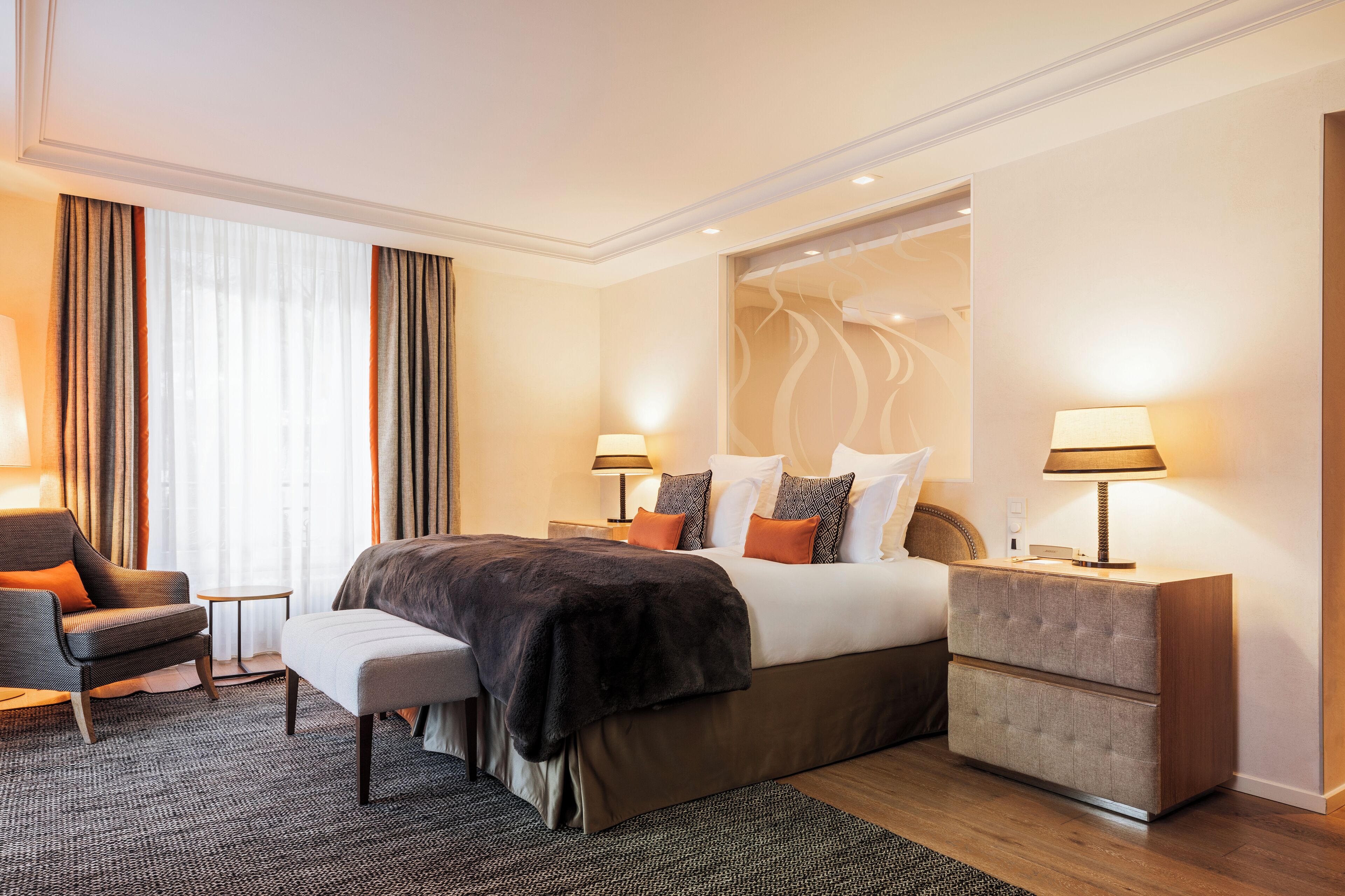 Una habitación de hotel con una cama grande, dos mesitas de noche, una silla, un banco y vistas al exterior a través de amplios ventanales.