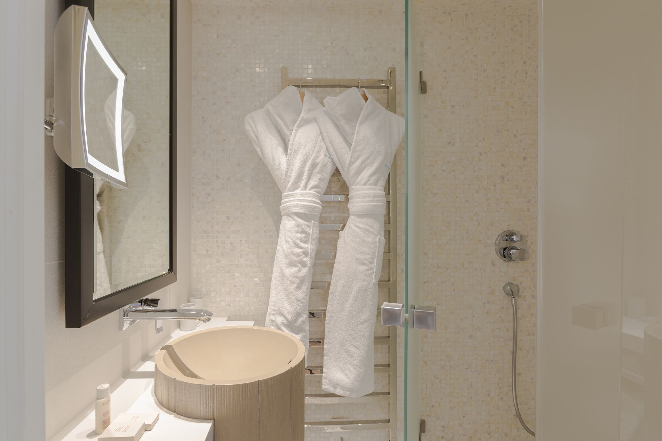 Ein Badezimmer mit einem runden Waschbecken, einem Spiegel, einer Duschkabine aus Glas und zwei weißen Handtüchern an einem Haken.