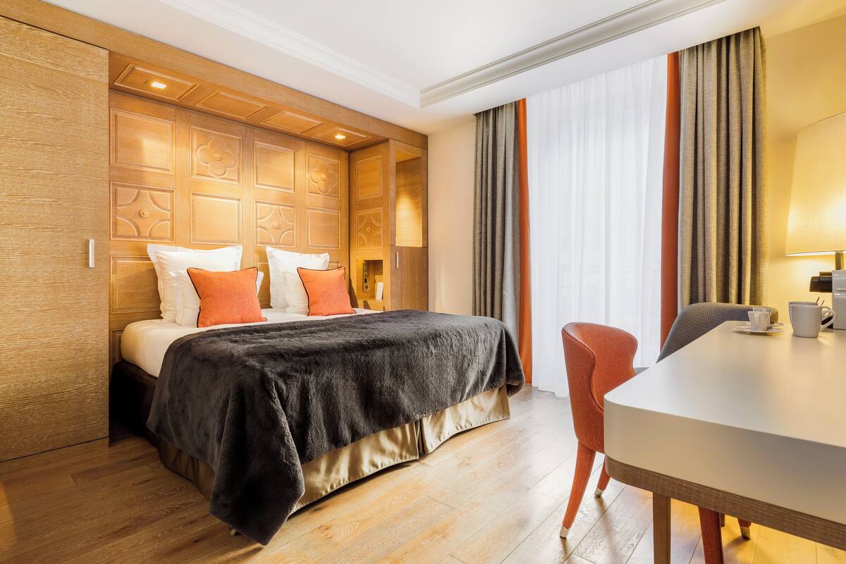 Une chambre d’hôtel avec une tête de lit en bois, un lit avec des oreillers blancs et orange, une couvre-lit noire, une table à manger avec une chaise et une fenêtre avec des rideaux.