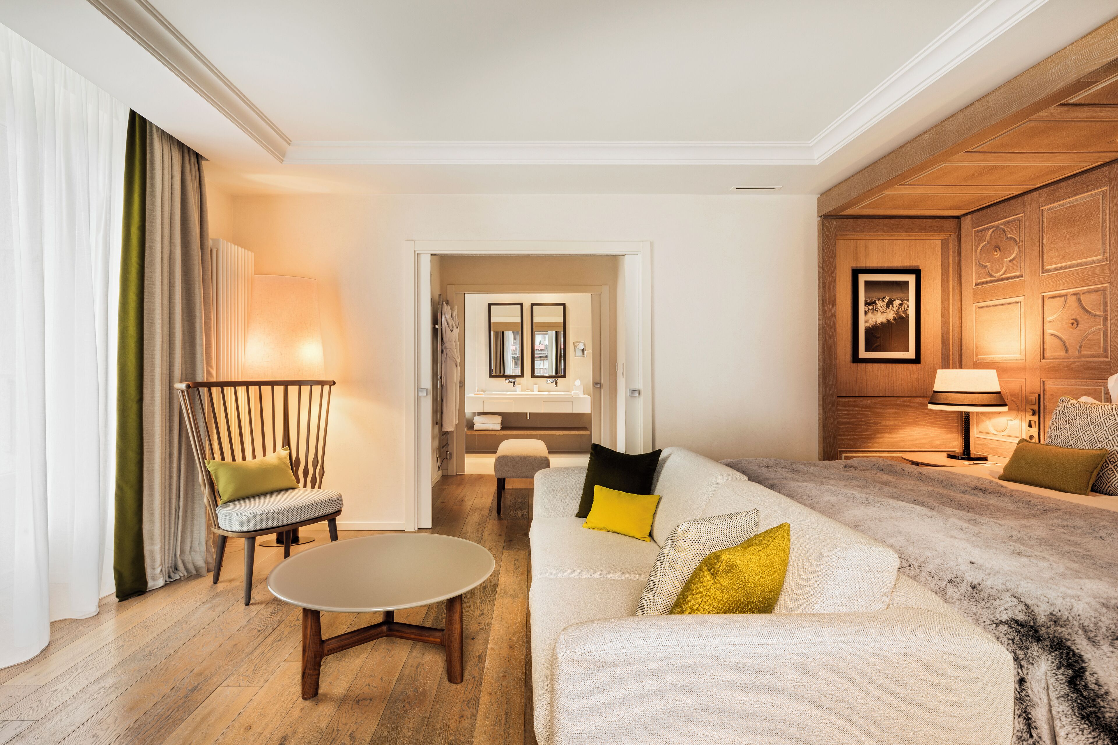 A modern hotel room with a wooden floor, a white sofa with yellow and black cushions, a wooden chair, a small round table, and a wooden cabinet with a framed picture.