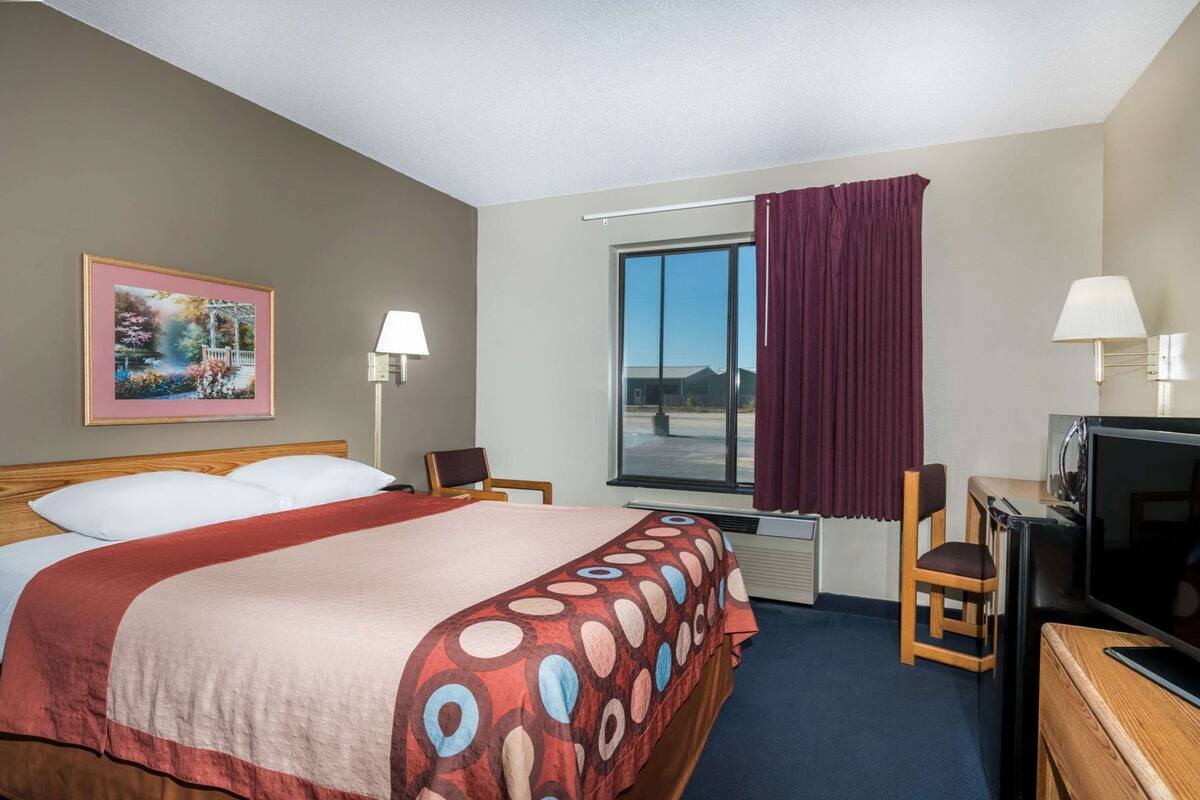 A hotel room with a bed, a desk with a television, a chair, a window with curtains, and a wall-mounted lamp.
