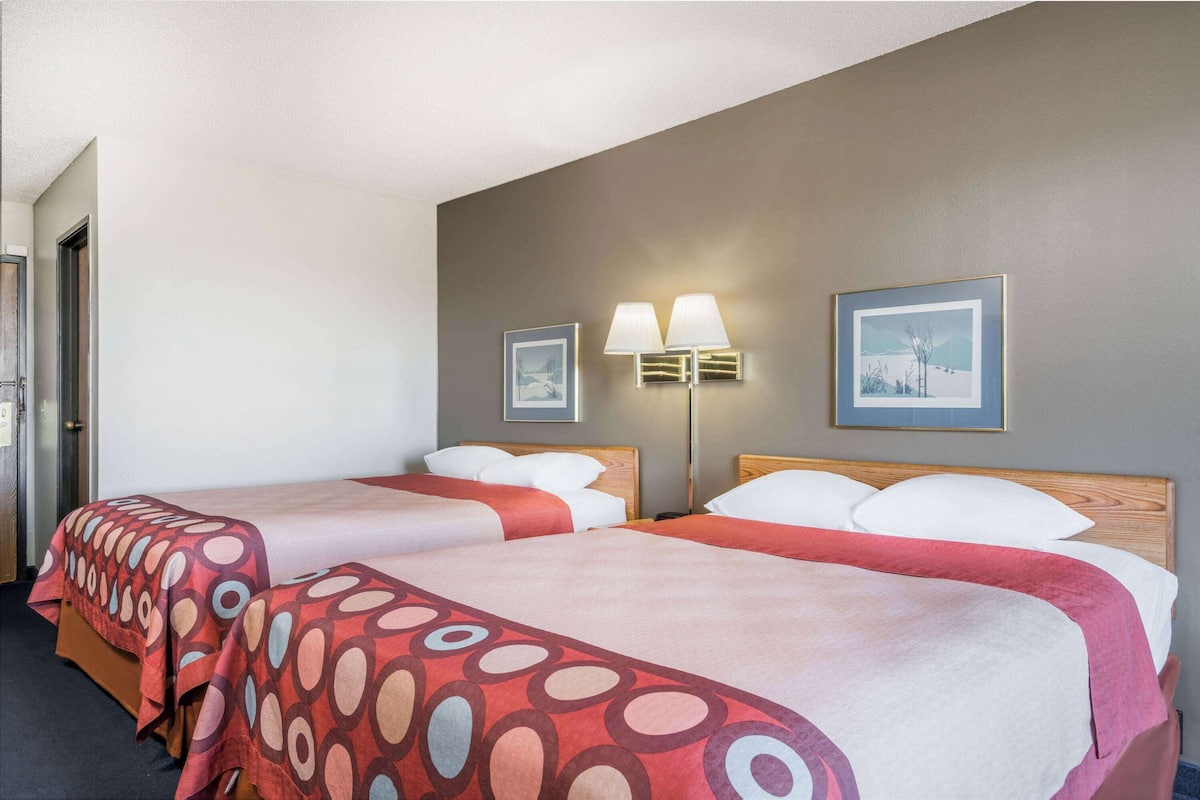A hotel room with two beds, each with a red and white patterned bedspread, a wooden headboard, and a nightstand with a lamp.
