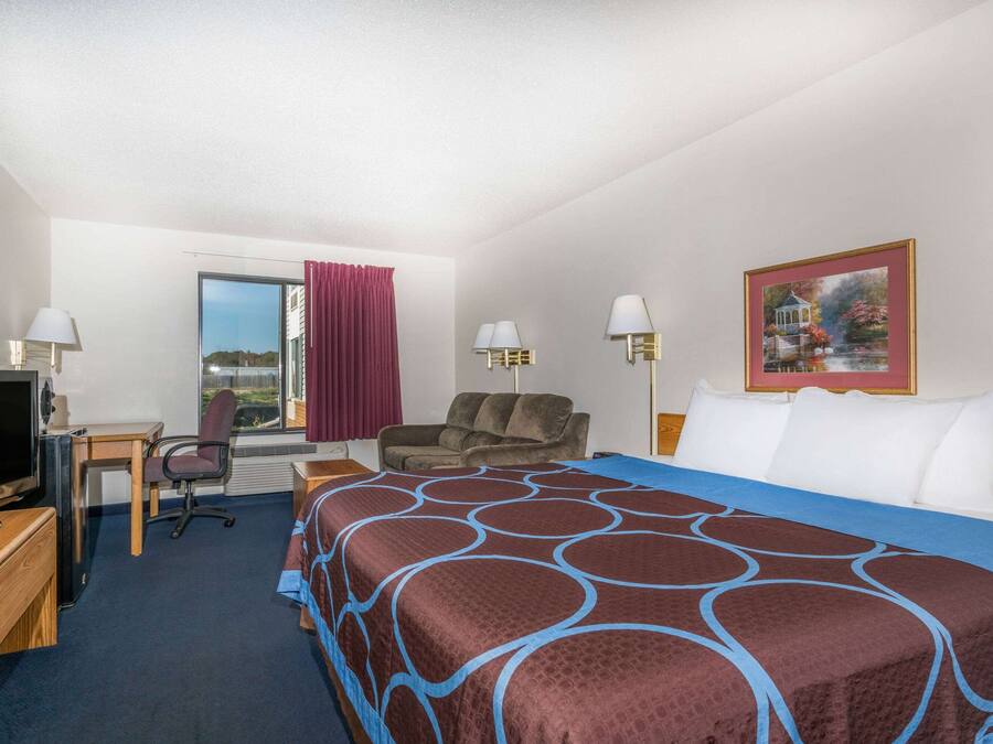 Ein Hotelzimmer mit einem Bett, einem Schreibtisch mit Fernseher, einem Stuhl, einem Fenster mit Vorhängen und einem Bild an der Wand.