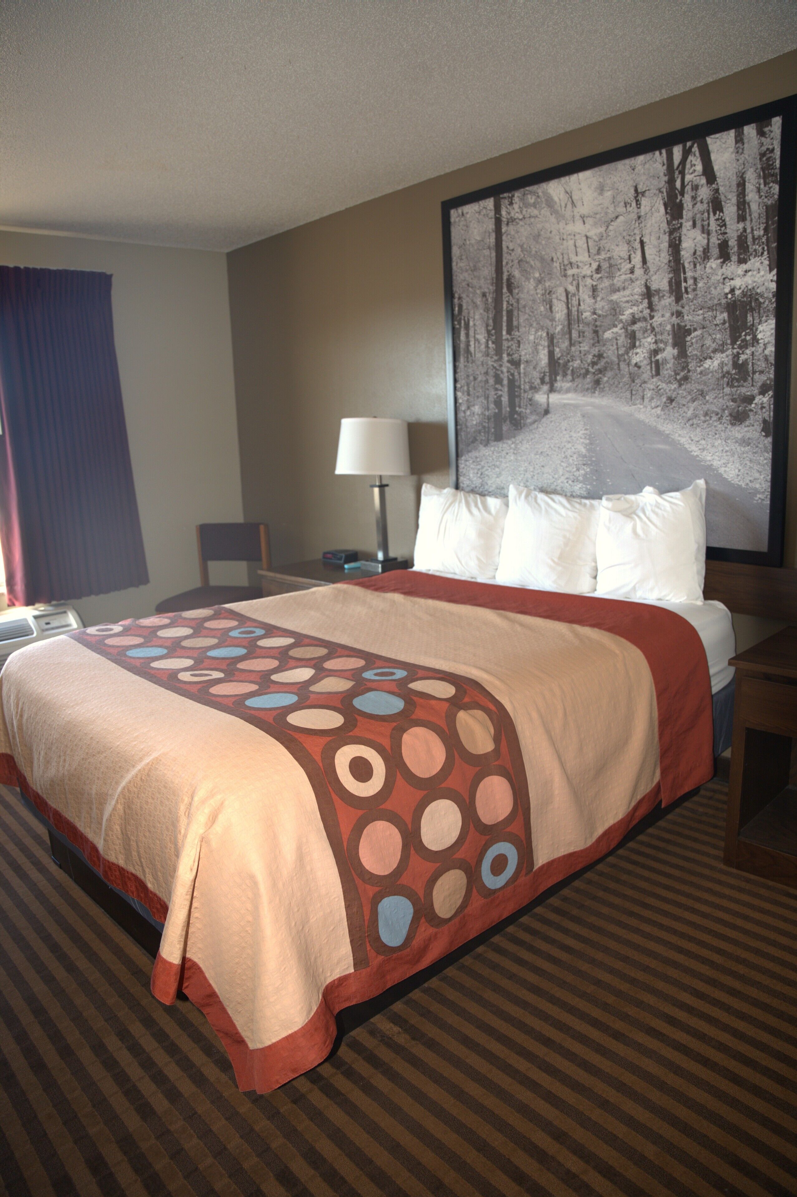 A hotel room with a large bed, a nightstand, a lamp, and a large framed picture of a forested road.