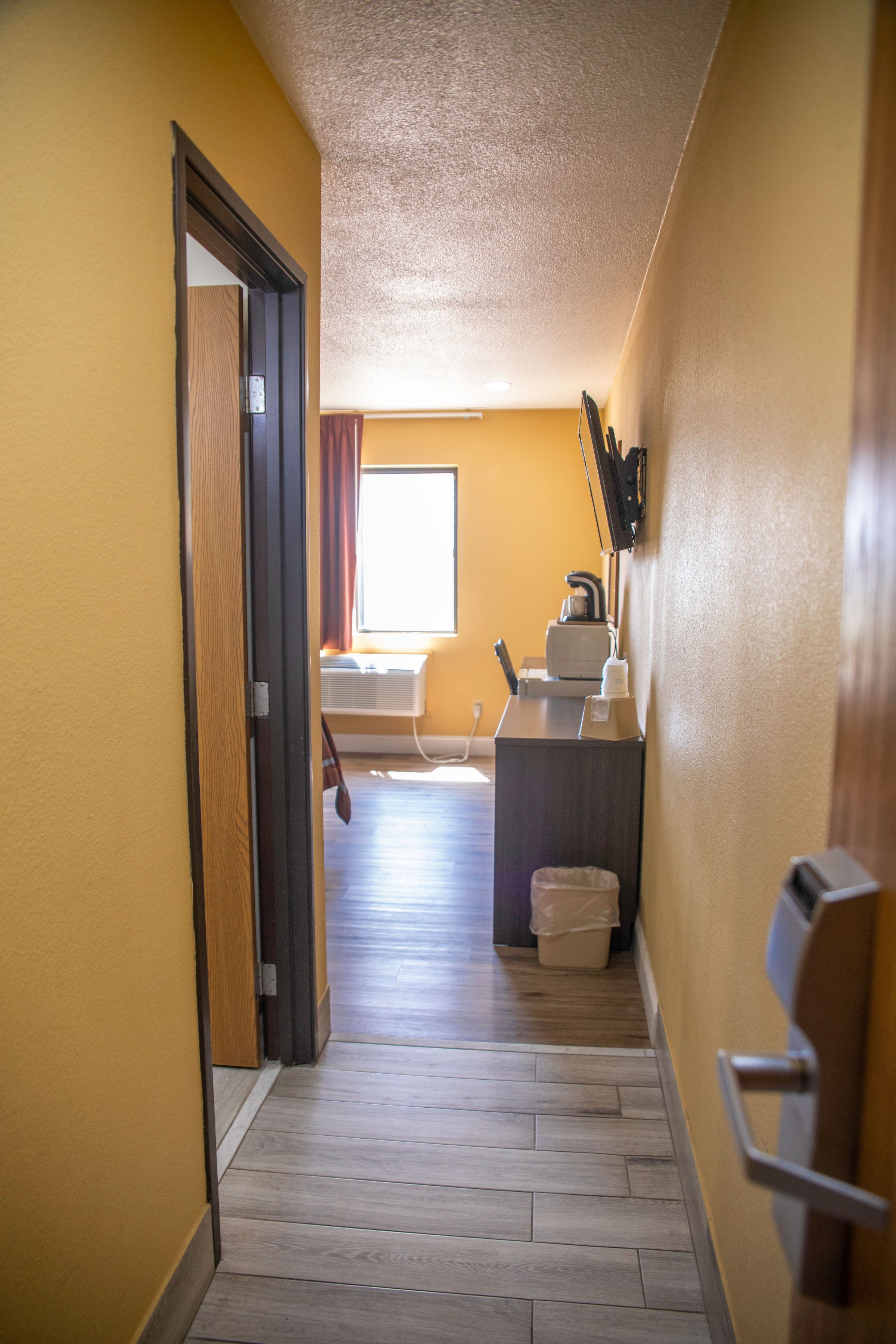 Un pasillo estrecho con suelo de madera, una puerta a la izquierda y una pequeña cocina con ventana y cortinas al fondo.