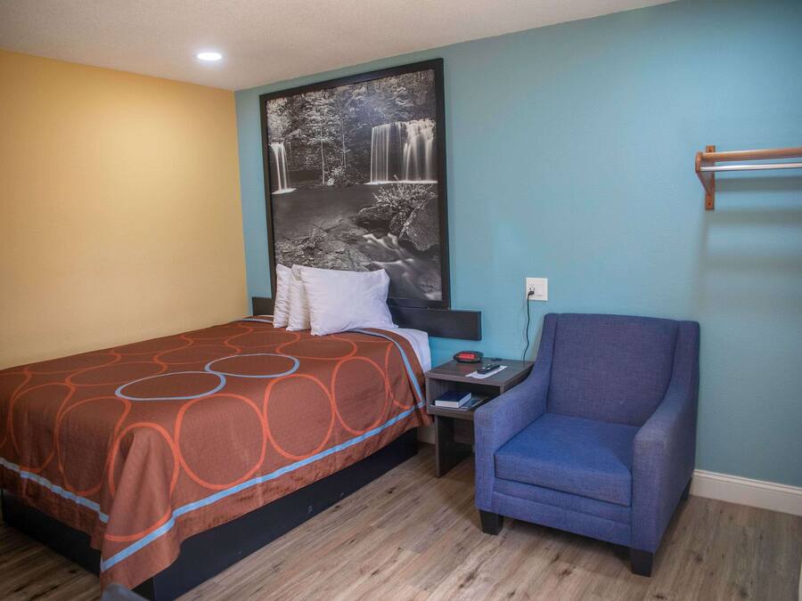 Quarto de hotel com cama, uma cadeira, mesa de cabeceira e prateleira fixada na parede.